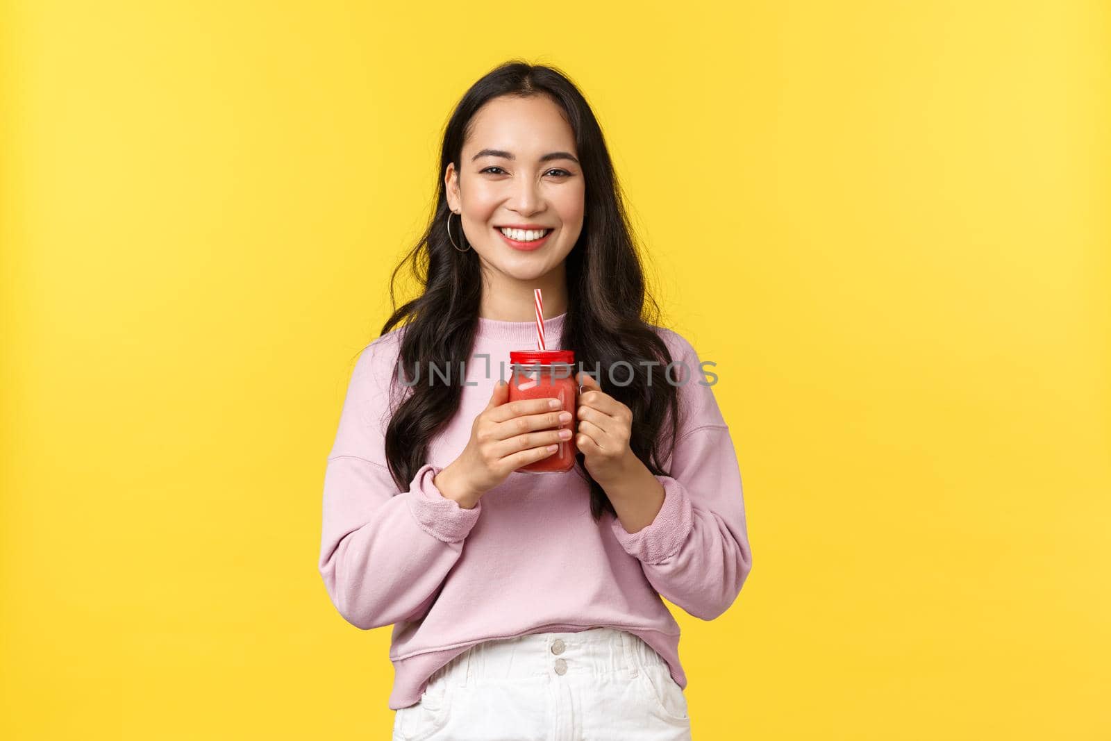 People emotions, lifestyle leisure and beauty concept. Cheerful smiling asian girl enjoying fresh smoothie, drinking juice and looking camera enthusiastic, standing yellow background.