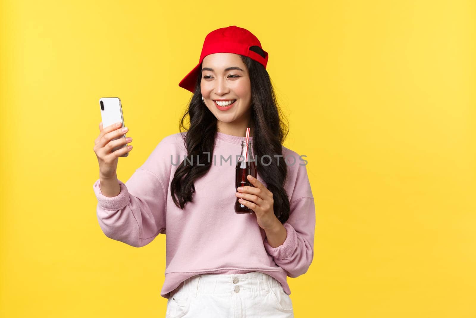 People emotions, drinks and summer leisure concept. Stylish cute asian female blogger in red cap, taking selfie with smartphone, drinking soda beverage and photographing herself by Benzoix