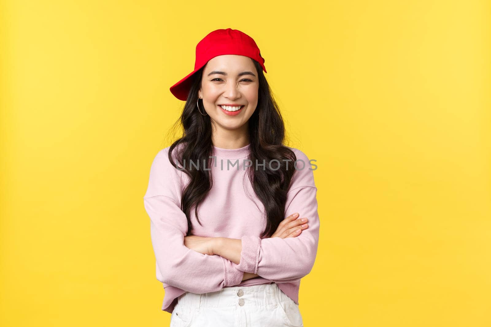 People emotions, lifestyle leisure and beauty concept. Happy cheerful asian woman smiling and laughing, cross arms chest, looking cool in hipster red cap over yellow background.