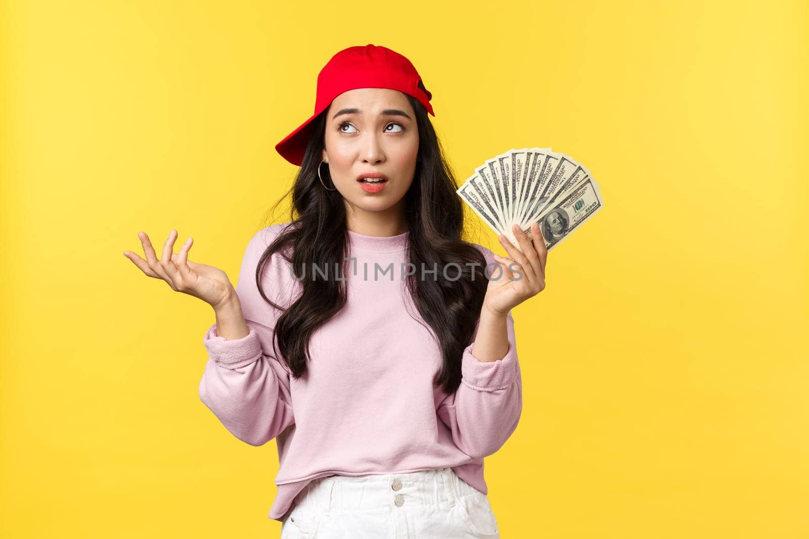 People emotions, lifestyle leisure and beauty concept. Unbothered cool and stylish rich girl in red cap, bragging about her wealth, showing money, careless spend cash, yellow background.