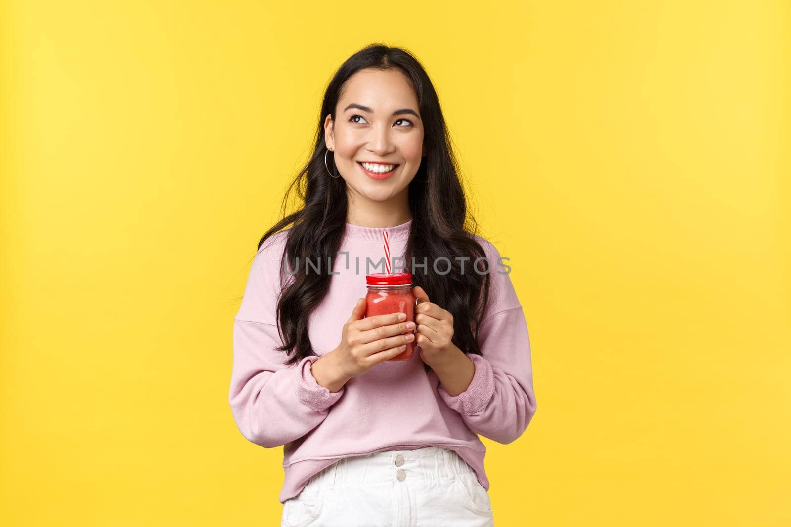 People emotions, lifestyle leisure and beauty concept. Smiling cute asian girl enjoying fresh cold smoothie on summer weekend, looking upper left corner dreamy, standing yellow background.