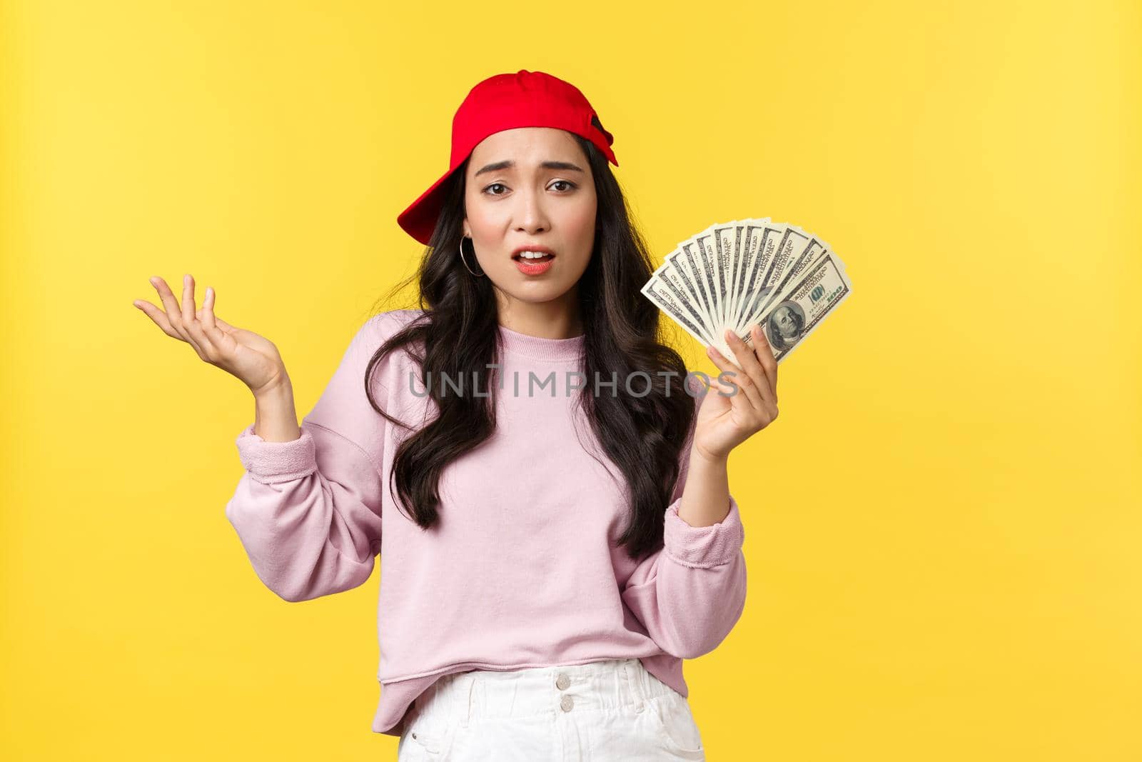 People emotions, lifestyle leisure and beauty concept. Unbothered cool and stylish rich girl in red cap, bragging about her wealth, showing money, careless spend cash, yellow background.