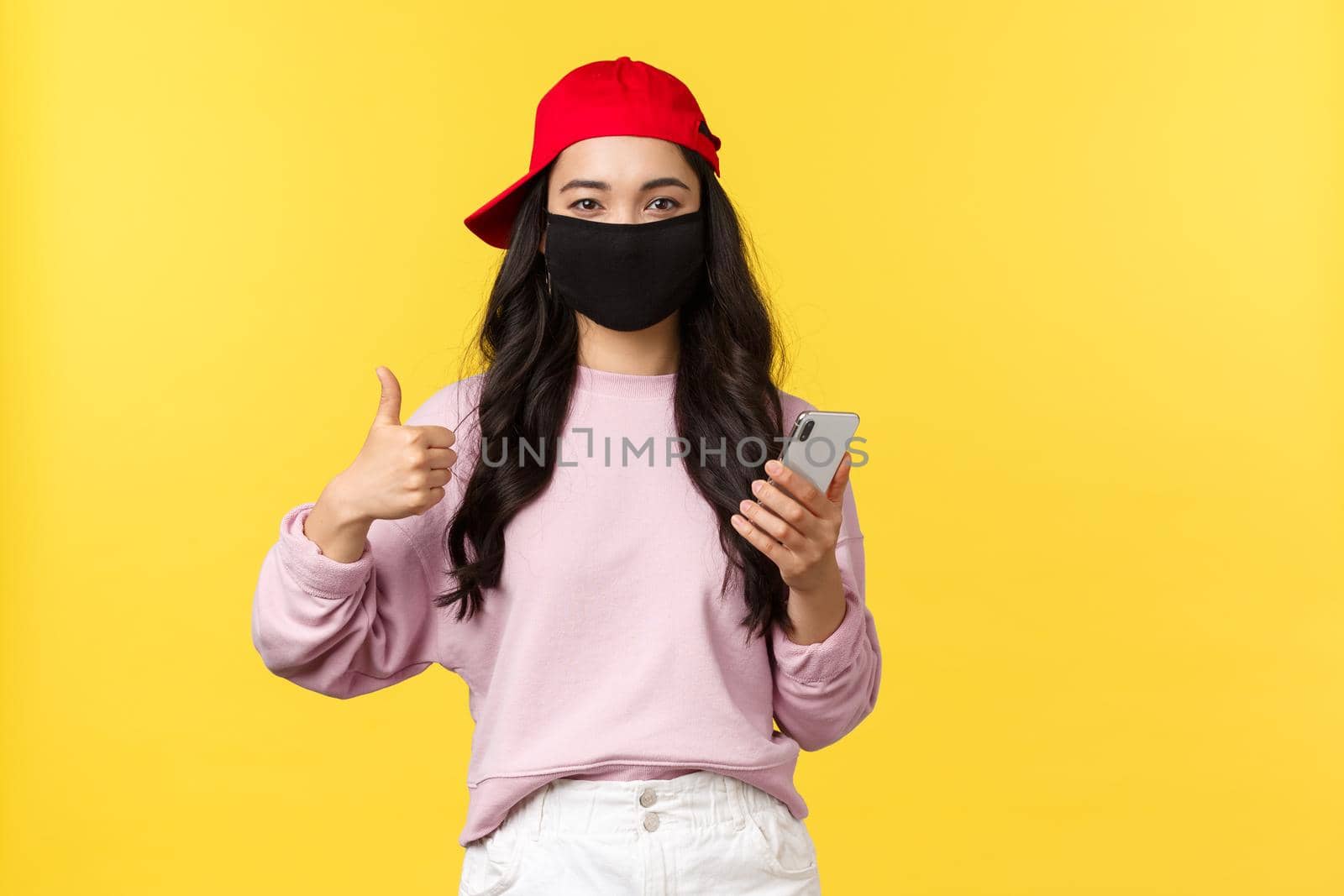 Covid-19, social-distancing lifestyle, prevent virus spread concept. Smiling cheerful asian girl in red cap and face mask, protect health from coronavirus, make thumbs-up, using mobile phone.