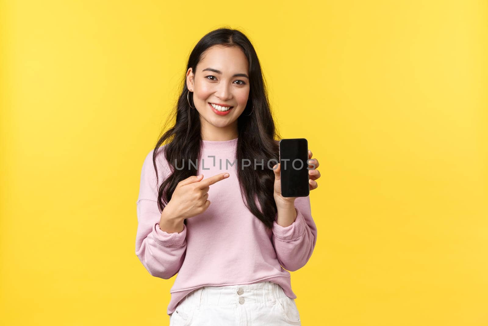 People emotions, lifestyle leisure and beauty concept. Smiling asian woman 20s, showing smartphone display, recommend application or mobile game, pointing finger at screen, yellow background by Benzoix