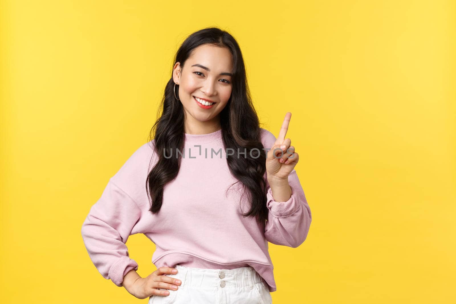 People emotions, lifestyle and fashion concept. Smiling happy good-looking asian girl giving advice, showing one finger as explain rules, make point, standing yellow background.