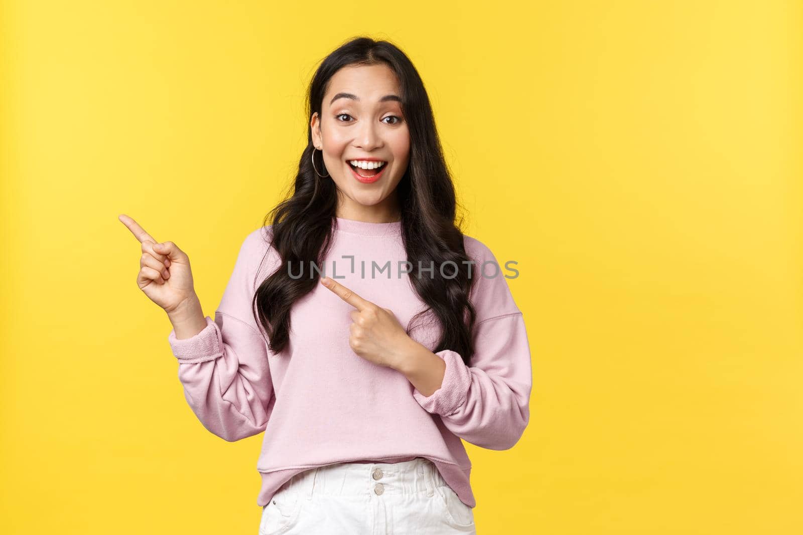 People emotions, lifestyle and fashion concept. Cheerful pretty korean girl in casual outfit, pointing upper left corner promo special discount banner or advertisement, stand over yellow background.