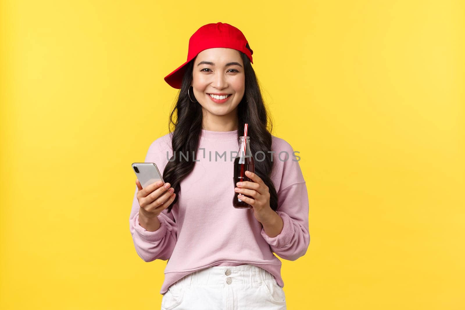 People emotions, drinks and summer leisure concept. Young teenage korean girl in red cap, messaging, using smartphone and drinking soda beverage, standing yellow background pleased.