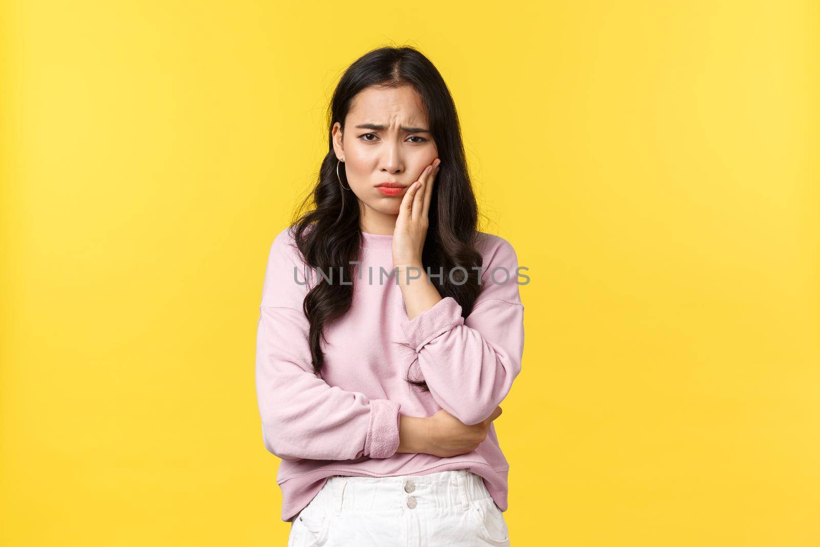 People emotions, lifestyle and fashion concept. Troubled uneasy young asian woman feeling toothache, touching teeth and grimacing from pain, standing for appointment dentist, yellow background.