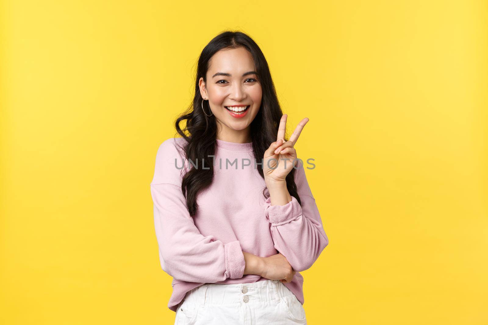 People emotions, lifestyle and fashion concept. Stylish korean girl in casual outfit showing peace sign and smiling silly, having good summer vacation, enjoying leisure, stand yellow background.