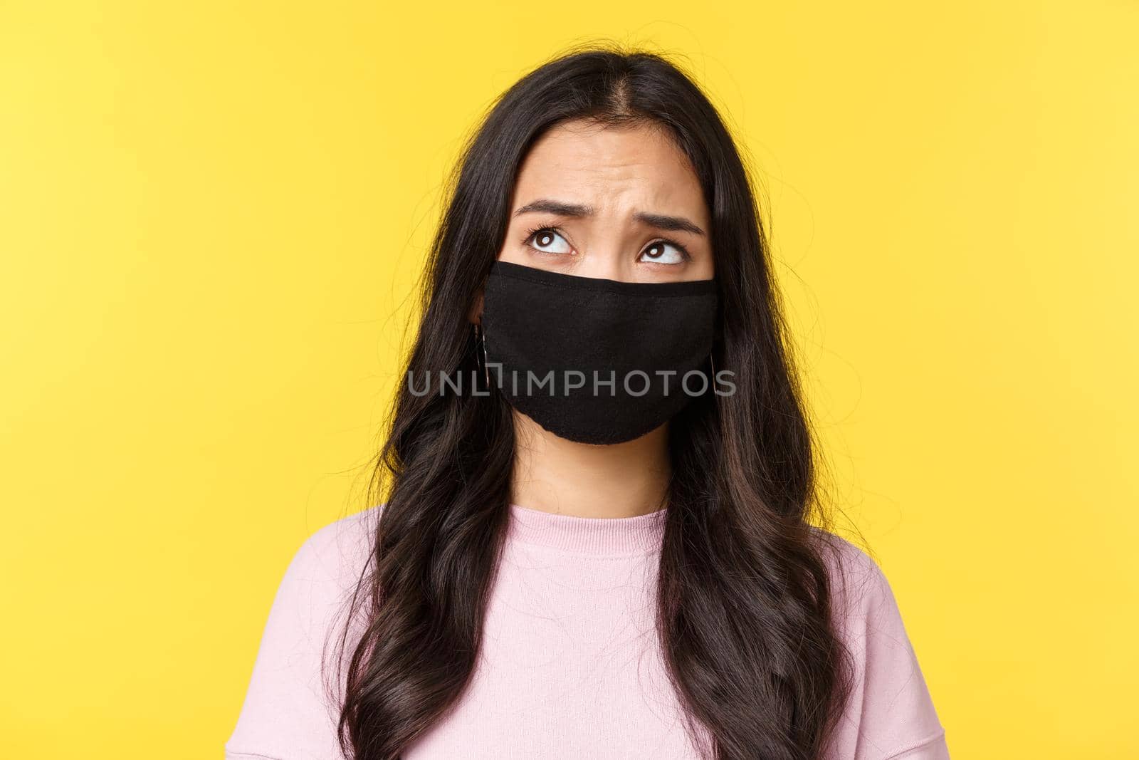 Covid-19, social-distancing lifestyle, prevent virus spread concept. Close-up of indecisive and concerned asian girl in face mask, looking top left corner hesitant, standing yellow background.