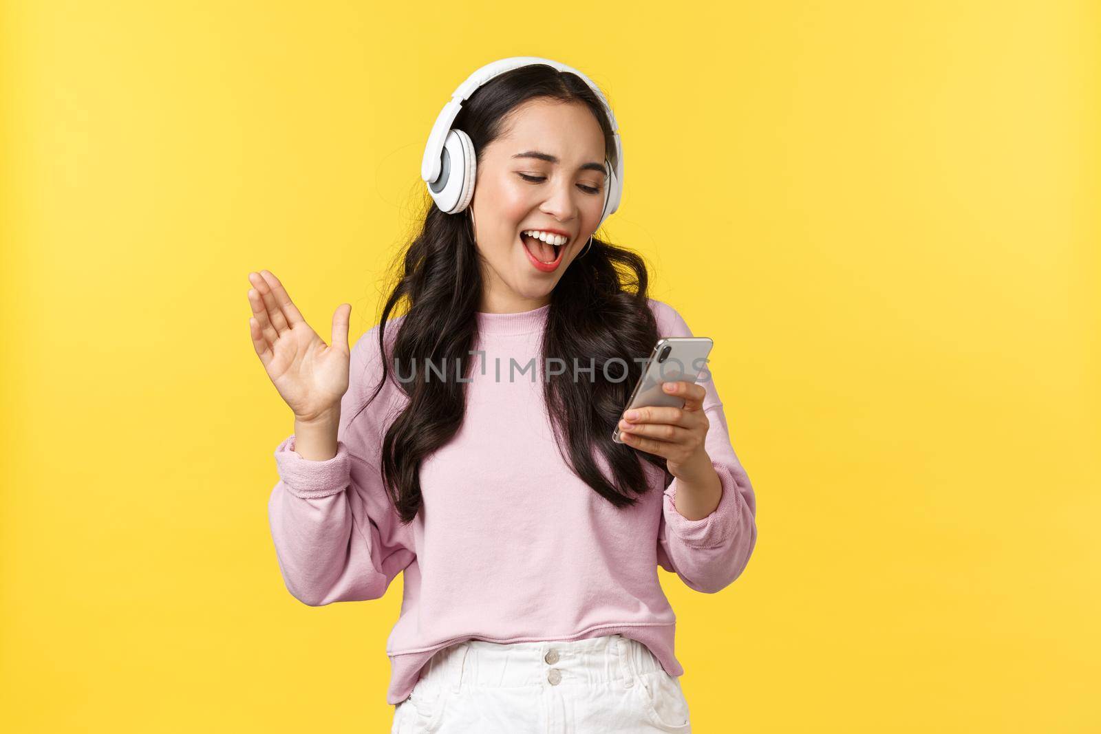 People emotions, lifestyle leisure and beauty concept. Smiling happy and carefree asian girl singing karaoke app in headphones, reading song from mobile phone, standing yellow background.