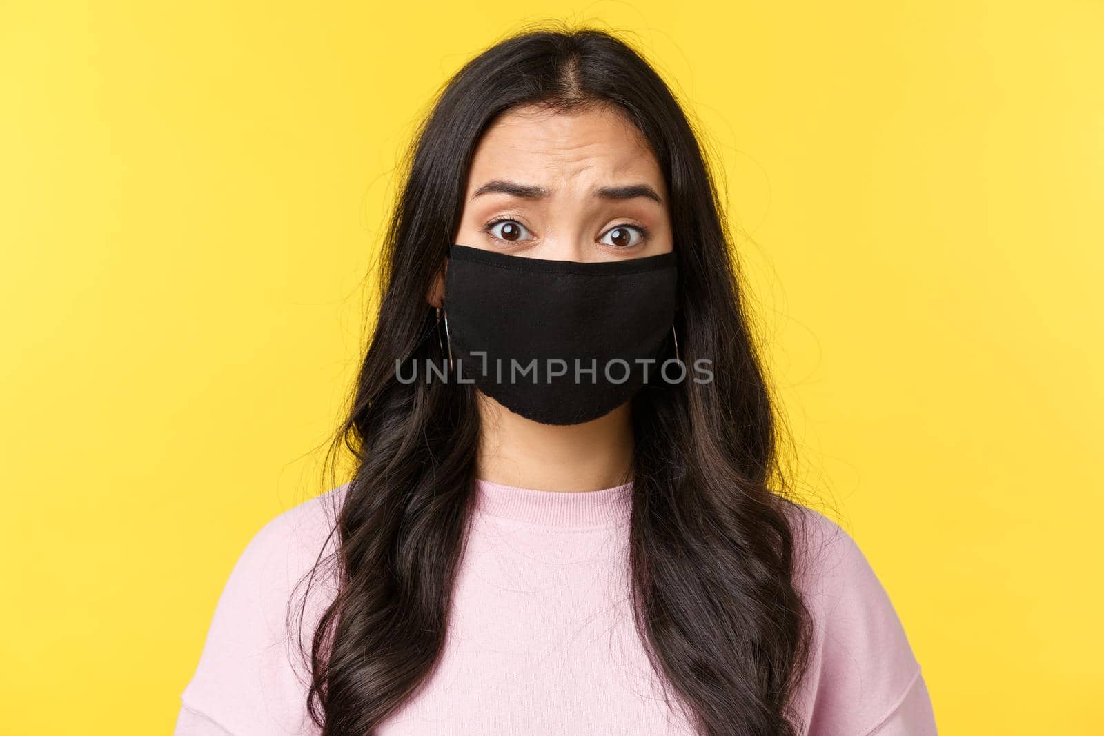 Covid-19, social-distancing lifestyle, prevent virus spread concept. Concerned and alarmed asian girl looking worried, wear face mask and frowning nervous, standing yellow background by Benzoix