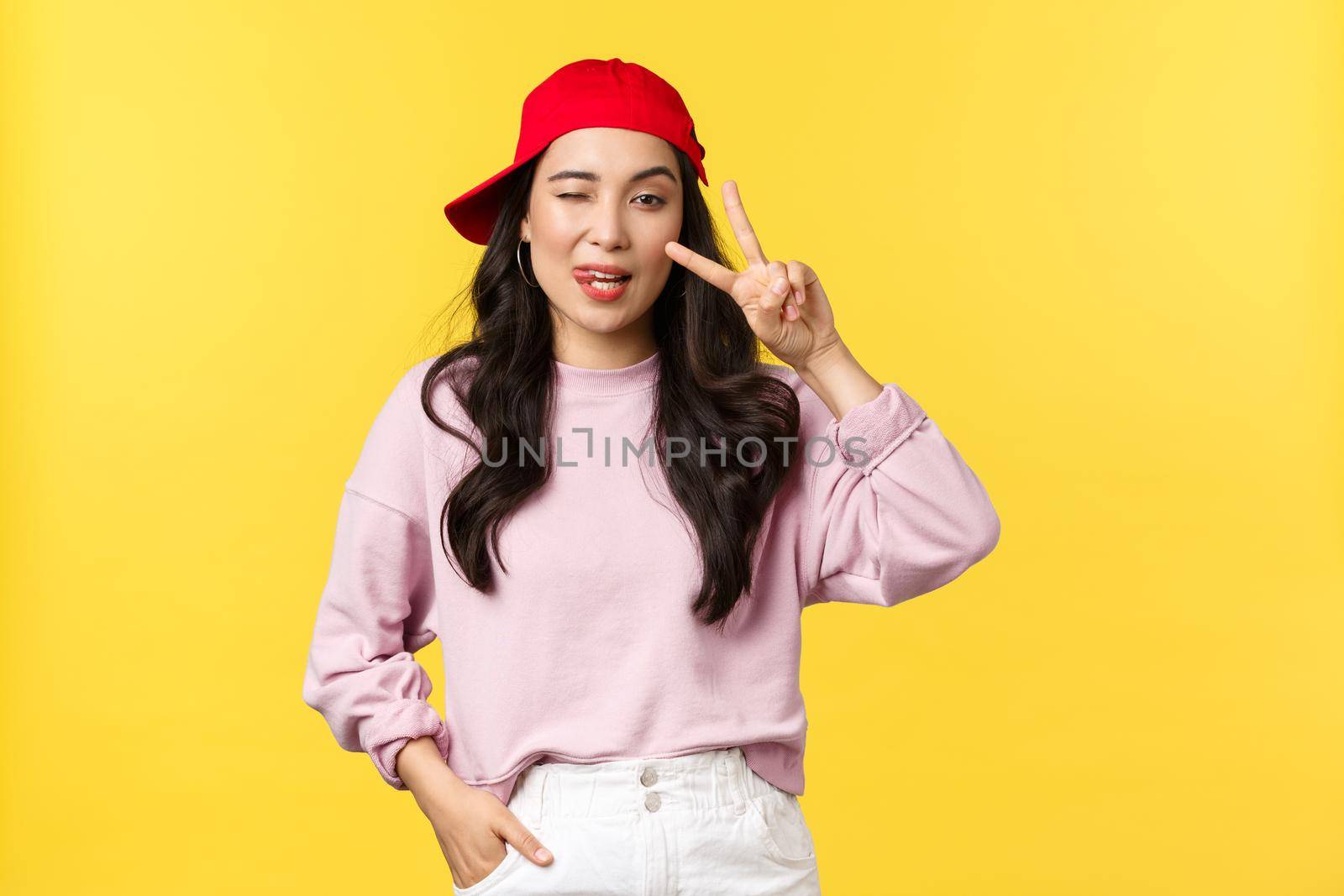 People emotions, lifestyle leisure and beauty concept. Stylish and cool asian girl in rnb red cap, showing peace sign and wink at camera, looking good in outfit over yellow background.