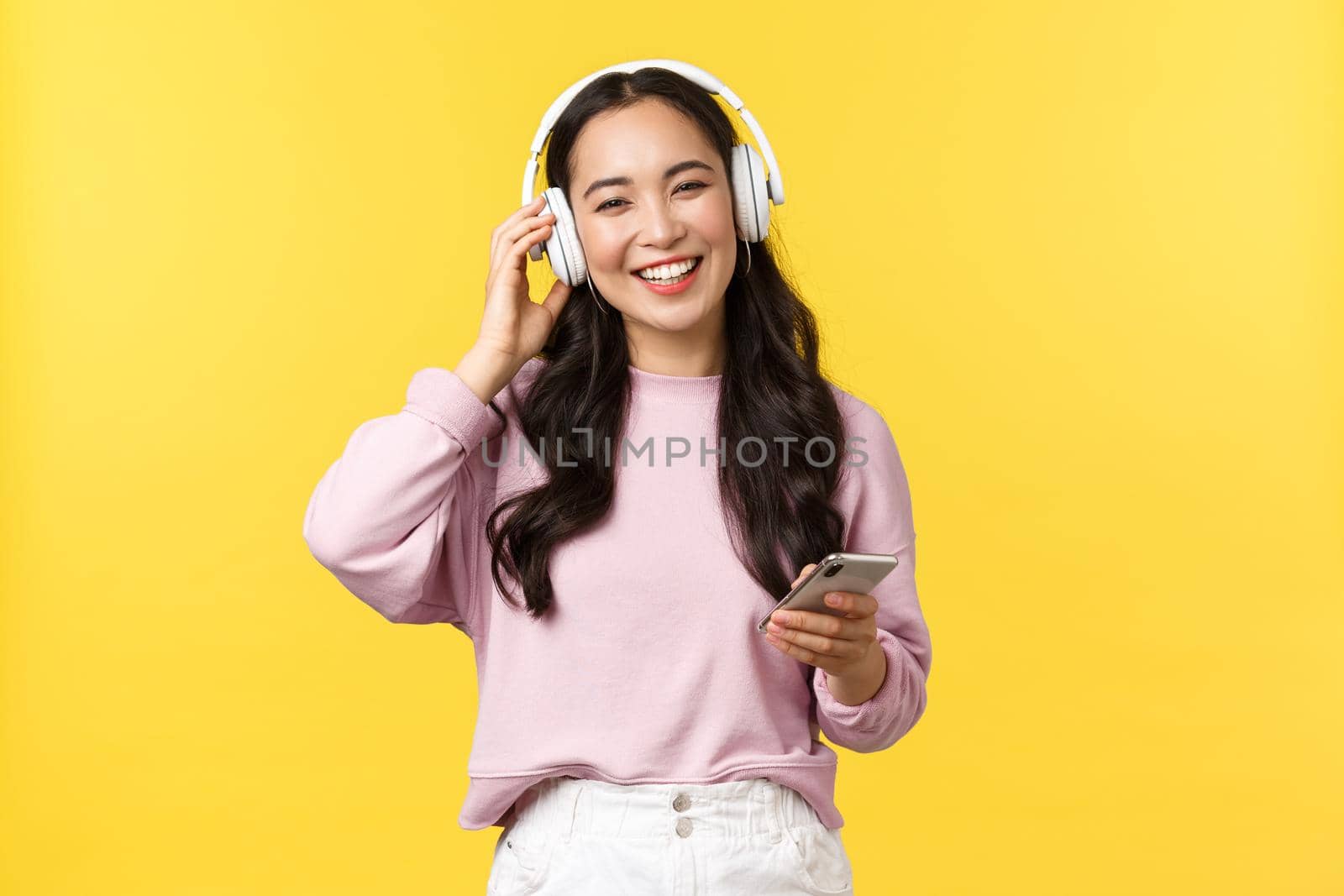 People emotions, lifestyle leisure and beauty concept. Happy smiling asian woman listening music in wireless headphones and looking pleased with nice sound, holding mobile phone by Benzoix
