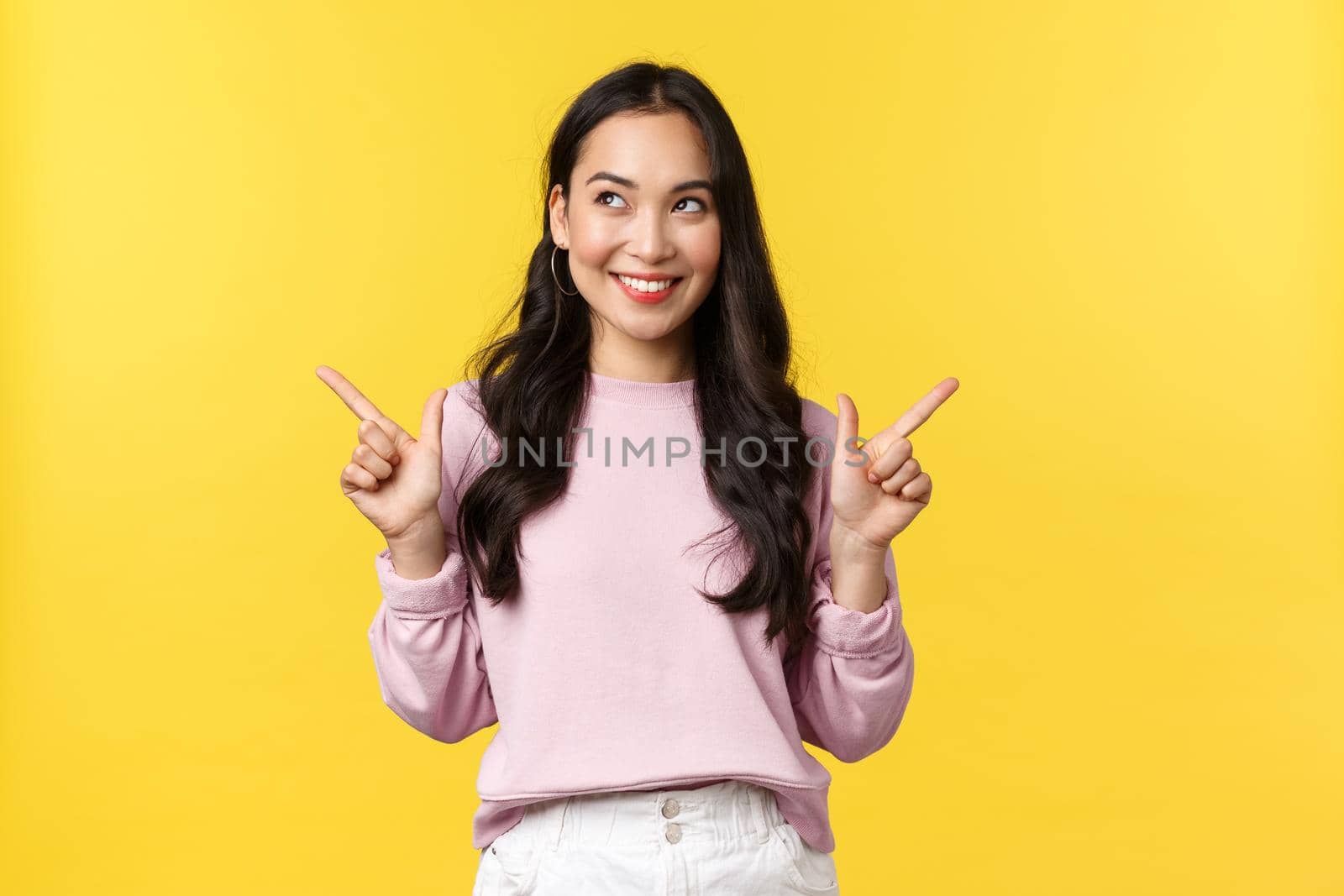 People emotions, lifestyle and fashion concept. Dreamy and thoughtful asian woman thinking, looking left pleased and pointing sideways at both variants, deciding what better, yellow background.