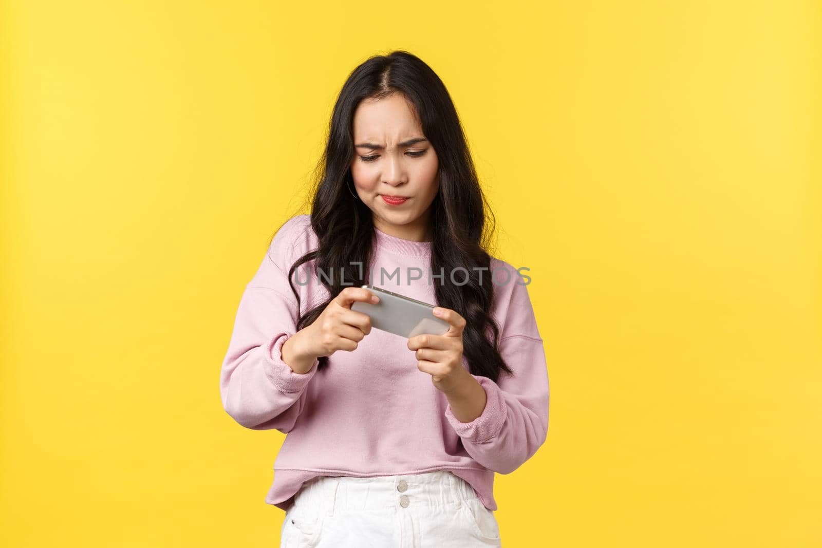 People emotions, lifestyle leisure and beauty concept. Focused and intense asian girl trying pass difficult level in mobile game, frowning as tap smartphone screen, standing yellow background.