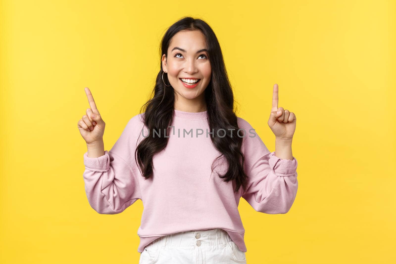 People emotions, lifestyle and fashion concept. Pleased smiling excited woman pointing fingers up and looking at top promo banner, something tasty or interesting, yellow background by Benzoix