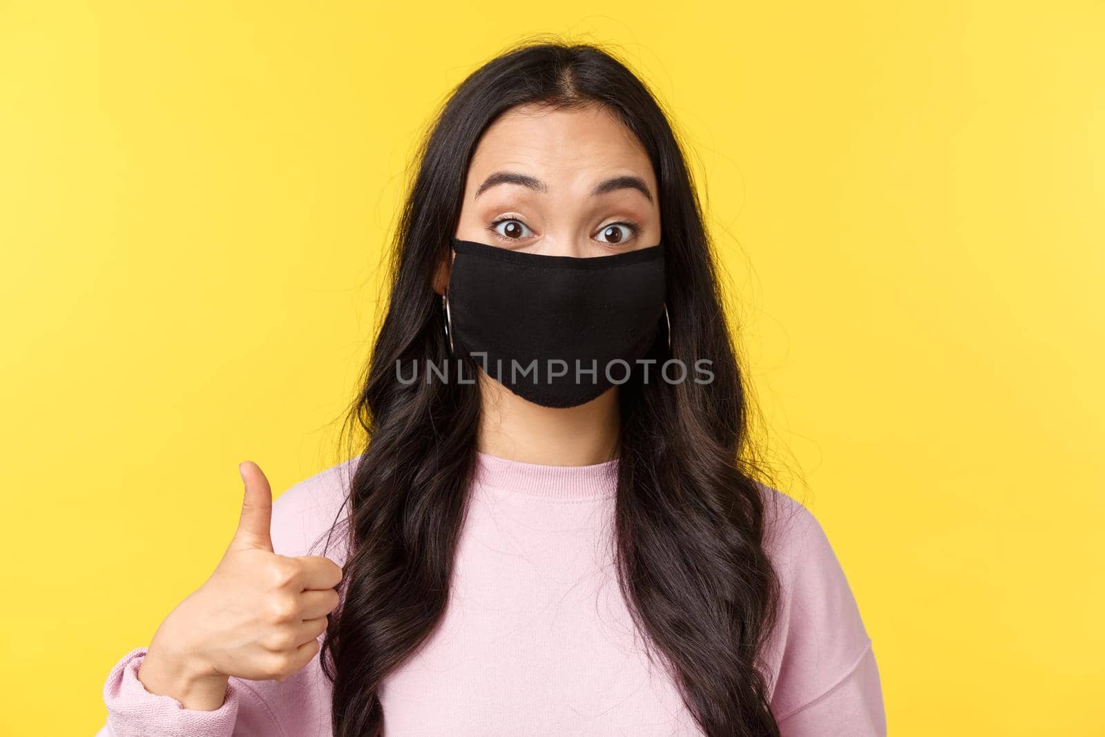 Covid-19, social-distancing lifestyle, prevent virus spread concept. Close-up of enthusiastic asian girl in face mask, look surprised and amused, showing thumbs-up in approval, yellow background.