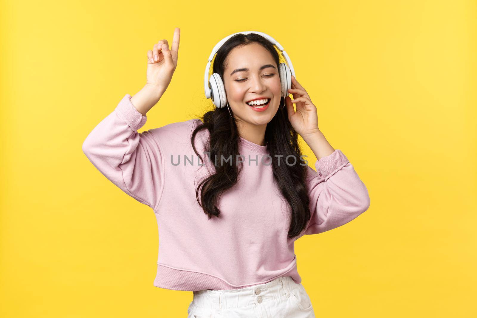 People emotions, lifestyle leisure and beauty concept. Pretty happy asian woman in wireless headphones, listening music and dancing, singing along, enjoying summer, stand yellow background.