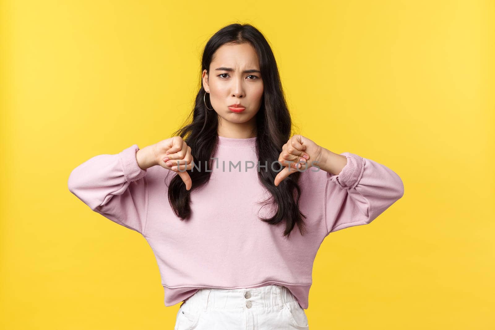 People emotions, lifestyle and fashion concept. Upset and disappointed asian girl pouting and showing disapproval, make thumbs-down in dislike, dont like idea, yellow background.