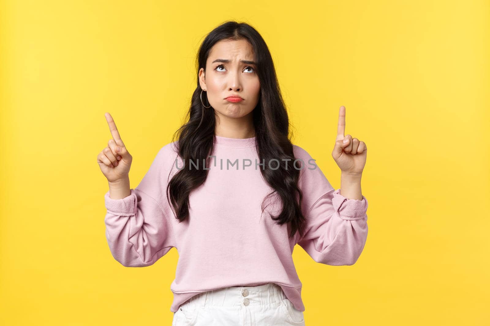 People emotions, lifestyle and fashion concept. Upset and gloomy pretty asian female express regret or disappointment, looking and pointing fingers up, grimacing dissatisfied, yellow background.
