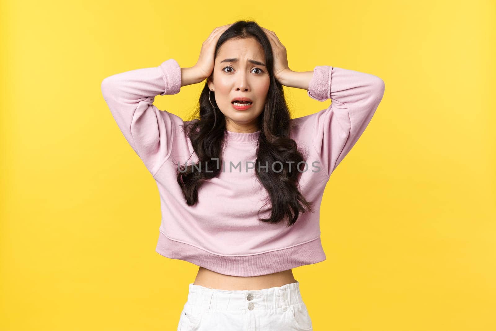 Lifestyle, emotions and advertisement concept. Anxious asian woman in panic look at disaster. grab head and looking frustrated and alarmed, feeling nervous, standing yellow background.