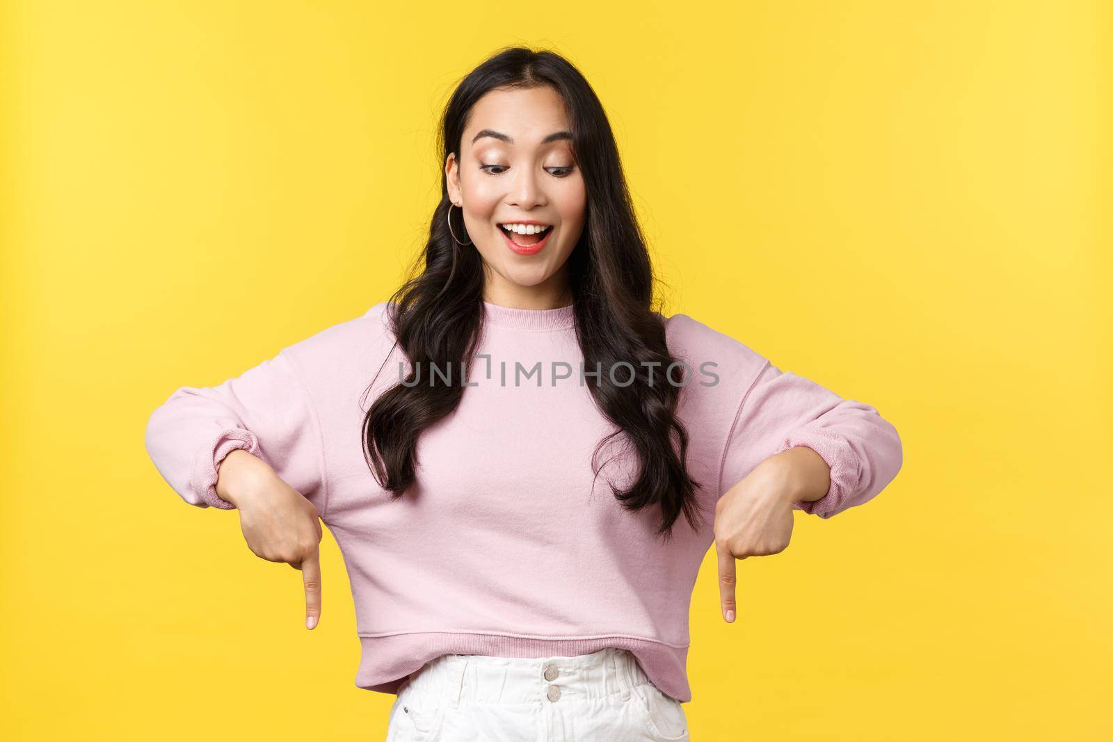 People emotions, lifestyle and fashion concept. Cheerful stylish asian girl pointing fingers down and looking at bottom advertisement with pleased smile, found perfect item on sale or shopping site.