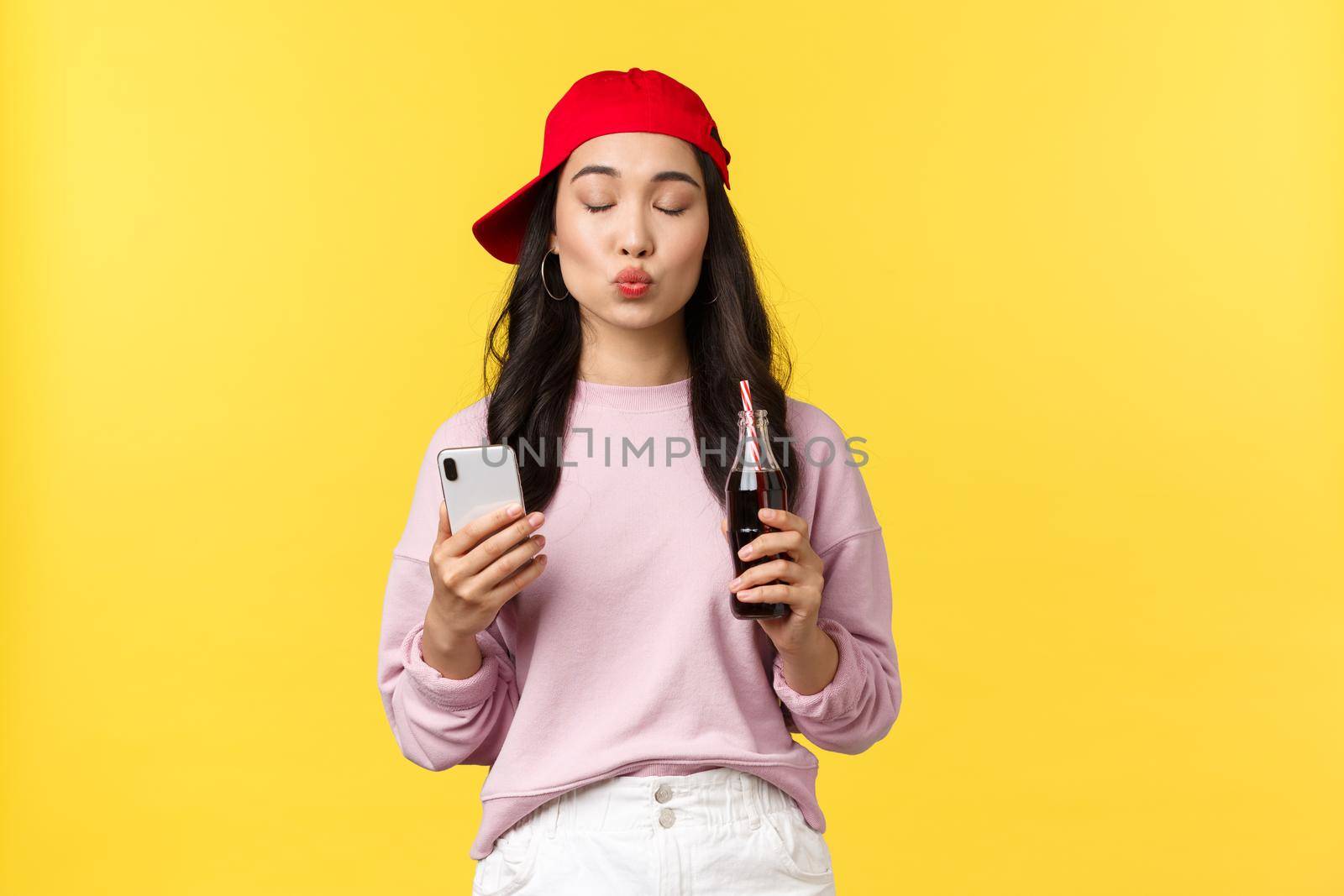 People emotions, drinks and summer leisure concept. Silly and cute asian girl in red cap, close eyes and fold lips to send air kiss, using smartphone and drinking soda from bottle, yellow background.