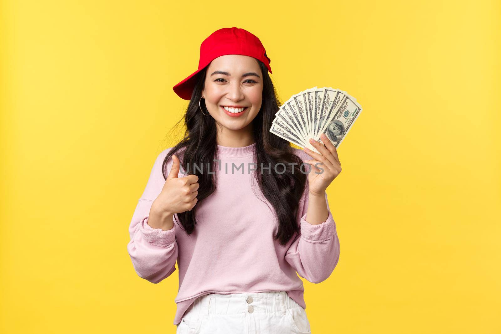 People emotions, lifestyle leisure and beauty concept. Cheerful and upbeat smiling korean girl in hip hop cap, showing thumbs-up and cash, explain how earn money on freelance, yellow background by Benzoix