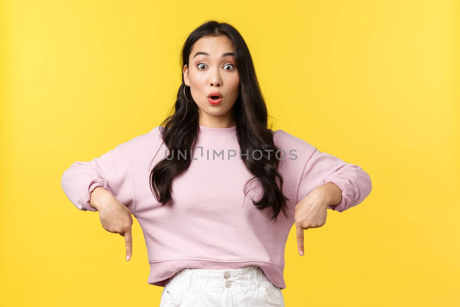 People emotions, lifestyle and fashion concept. Astonished and impressed pretty asian woman in stylish outfit pointing fingers down to show cool promo offer, advertise product over yellow background.