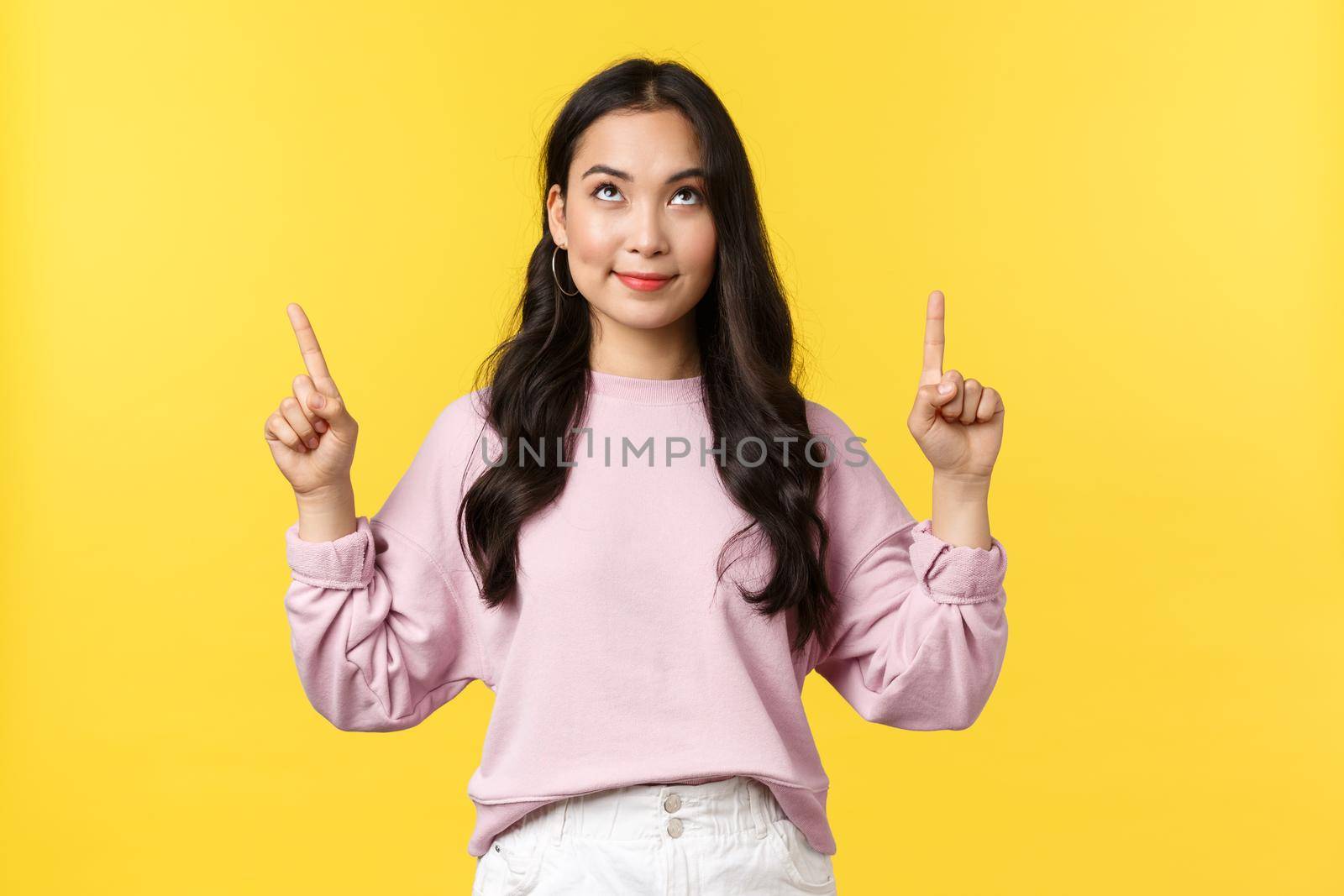 People emotions, lifestyle and fashion concept. Pleased dreamy pretty asian woman smiling and looking with temptation and desire at product upwards, pointing fingers up, yellow background.