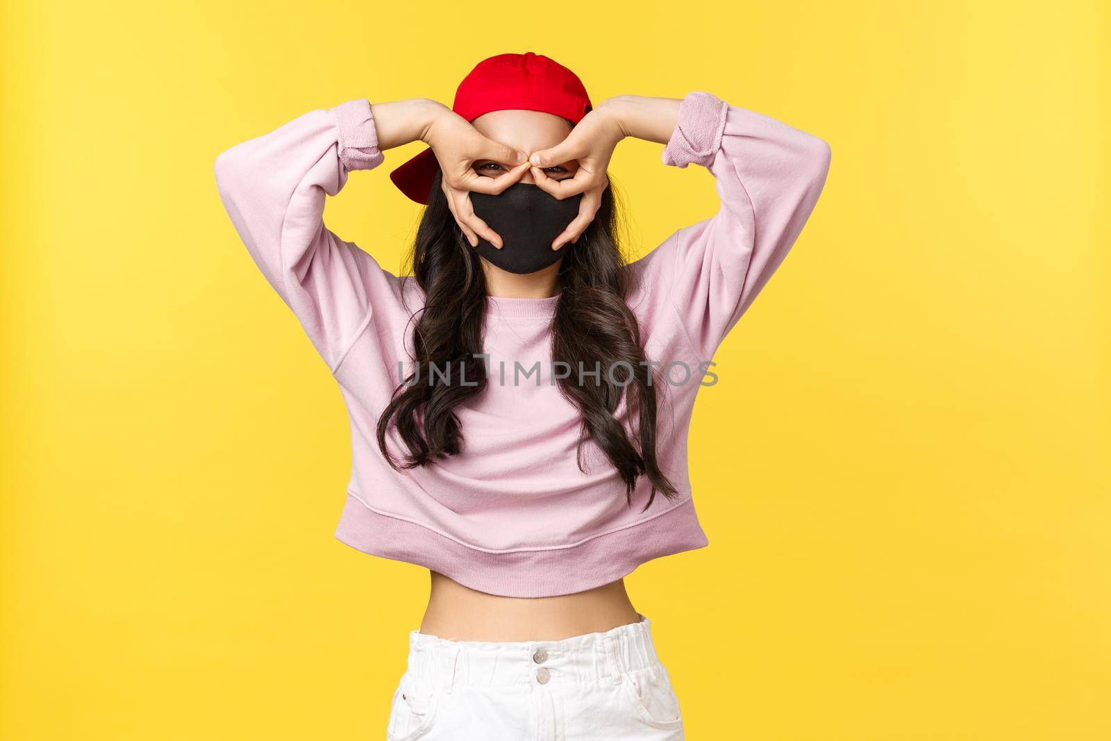 Covid-19, social-distancing lifestyle, prevent virus spread concept. Funny and cute asian girl in face mask and red cap, make fake glasses with fingers over eyes, stare surprised and impressed by Benzoix