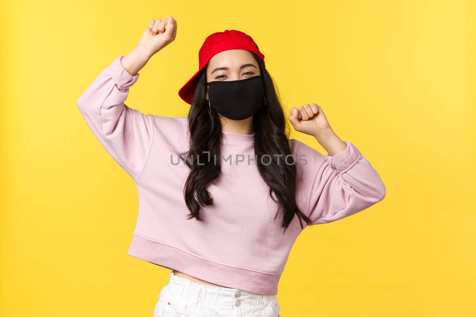 Covid-19, social-distancing lifestyle, prevent virus spread concept. Carefree excited asian girl in red cap and face mask, dancing with hands raised up, having good summertime, yellow background.