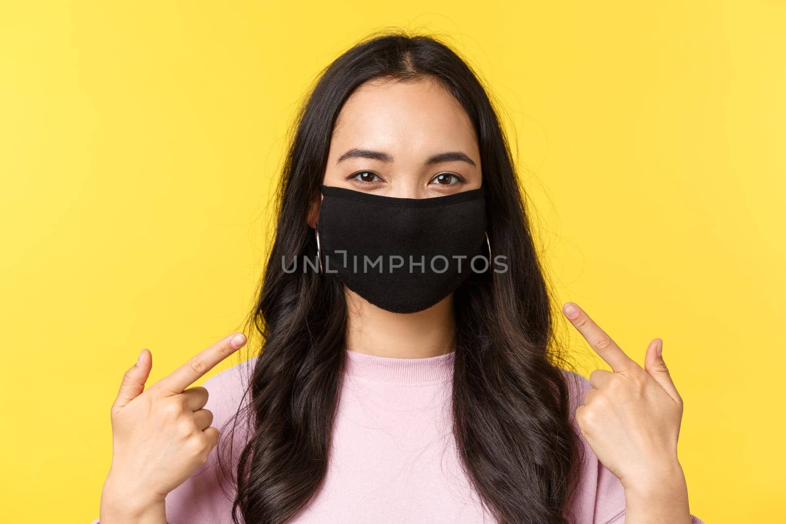 Covid-19, social-distancing lifestyle, prevent virus spread concept. Asian cheerful girl, smiling and pointing at face mask, recommend wearing protective measures during coronavirus pandemic.