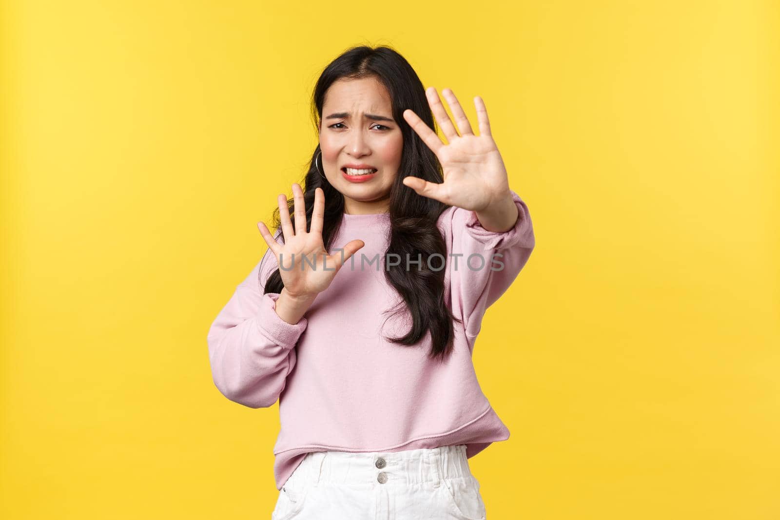 People emotions, lifestyle and fashion concept. Alarmed and displeased asian woman raising hands defensive, asking to stop, feeling anxious and scared standing yellow background.