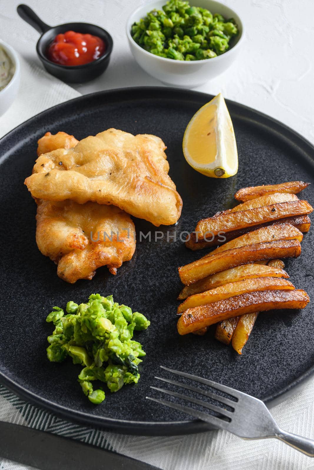 British Traditional Fish and chips with minty mashed peas detail and a slice of lemon. on black round plate over white lintn and stone surface by Ilianesolenyi