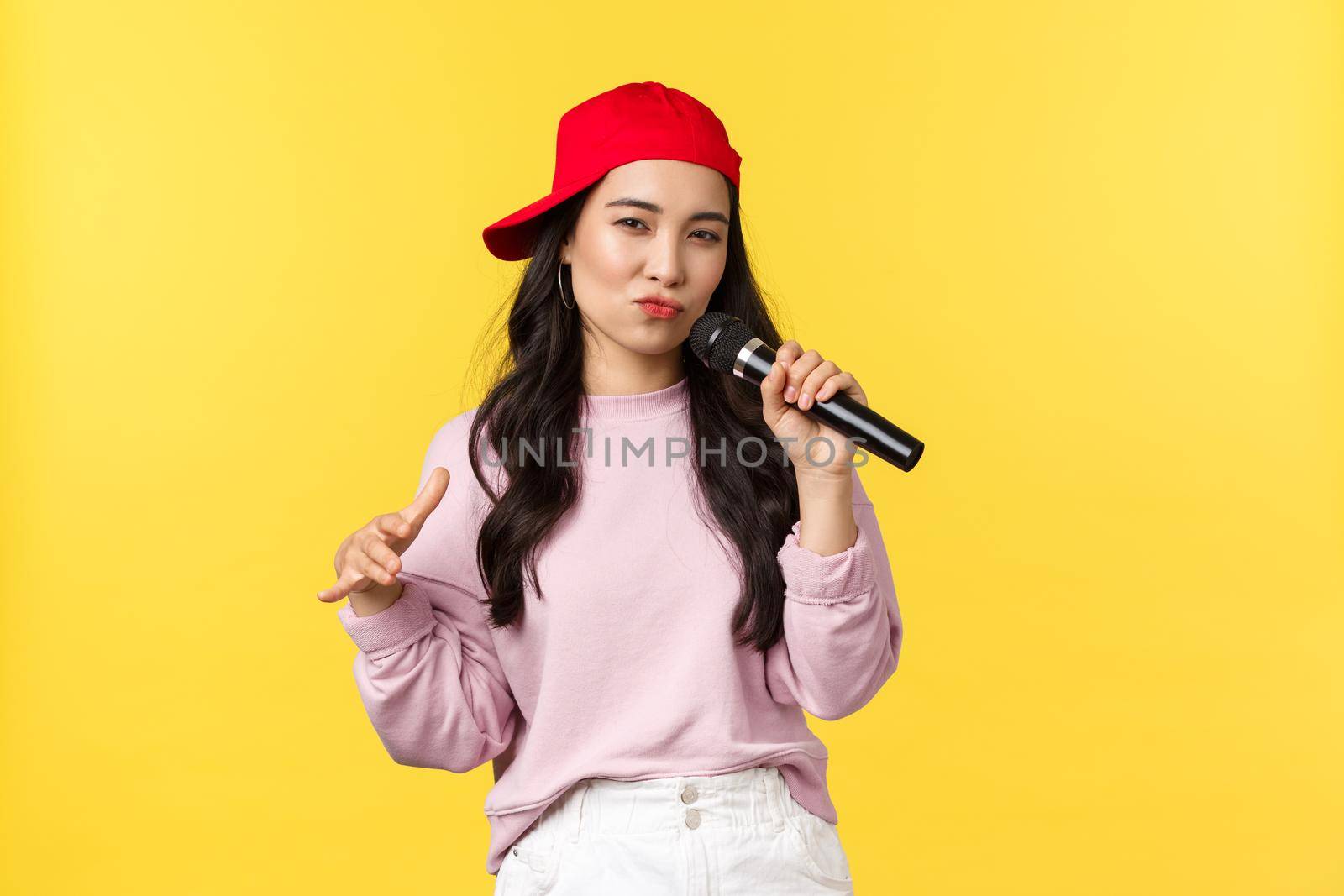 People emotions, lifestyle leisure and beauty concept. Carefree emotive young asian female hip hop singer in red cap, singing song in microphone with passion, yellow background.