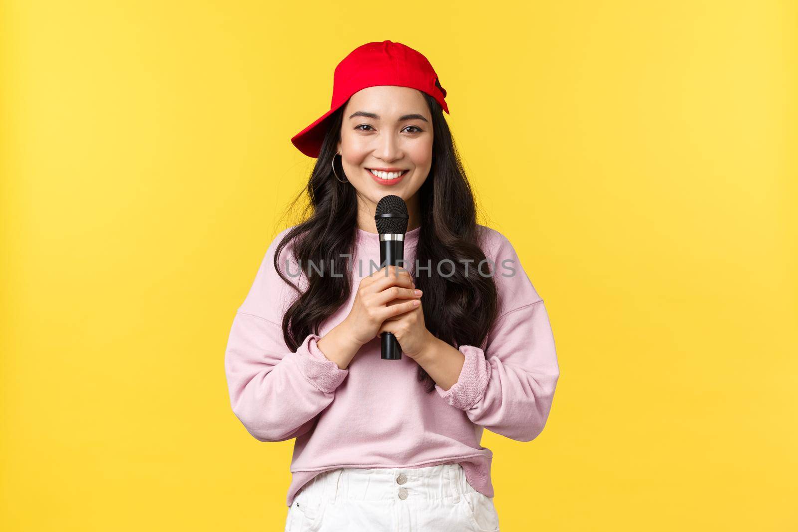 People emotions, lifestyle leisure and beauty concept. Cute smiling asian woman performer singing song in red cap, holding microphone as giving speech or sing rap, standing yellow background.
