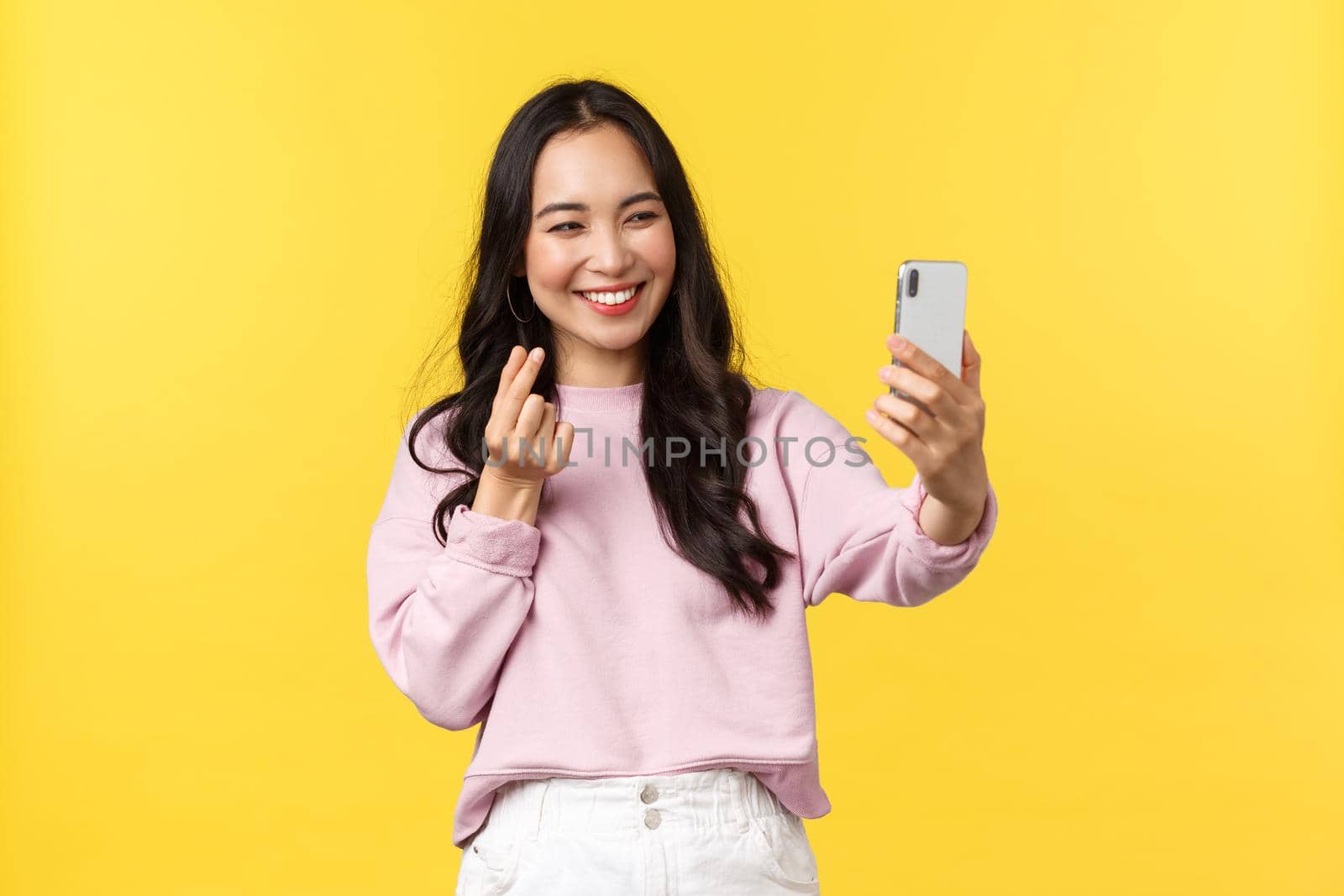 People emotions, lifestyle leisure and beauty concept. Cheerful asian girl over yellow background taking selfie on mobile phone, use photo filters app and show heart gesture.