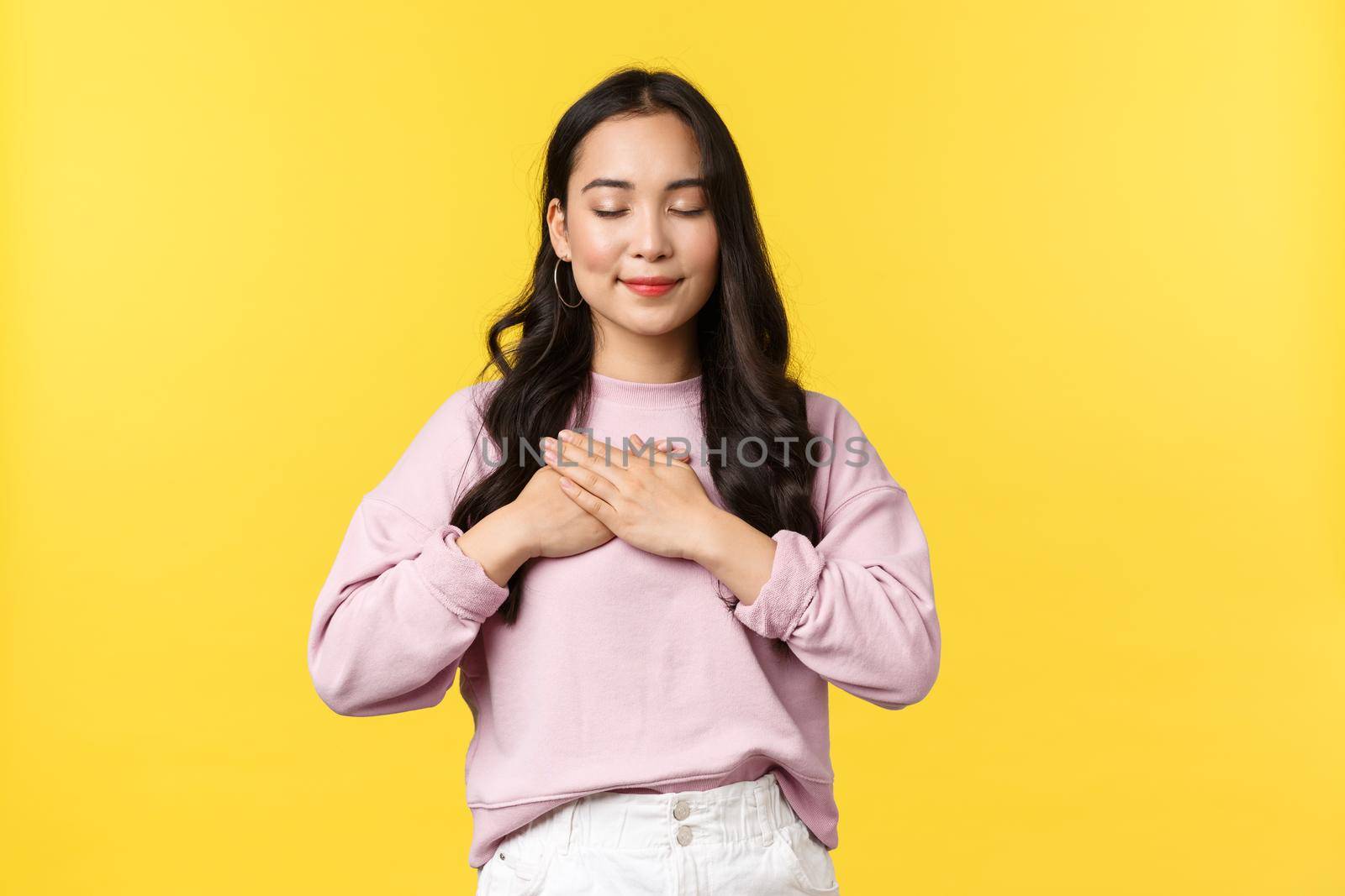 People emotions, lifestyle and fashion concept. Dreamy tender asian woman in casual outfit, close eyes and smiling, hold hands on heart, have romantic lovely memory, standing yellow background by Benzoix