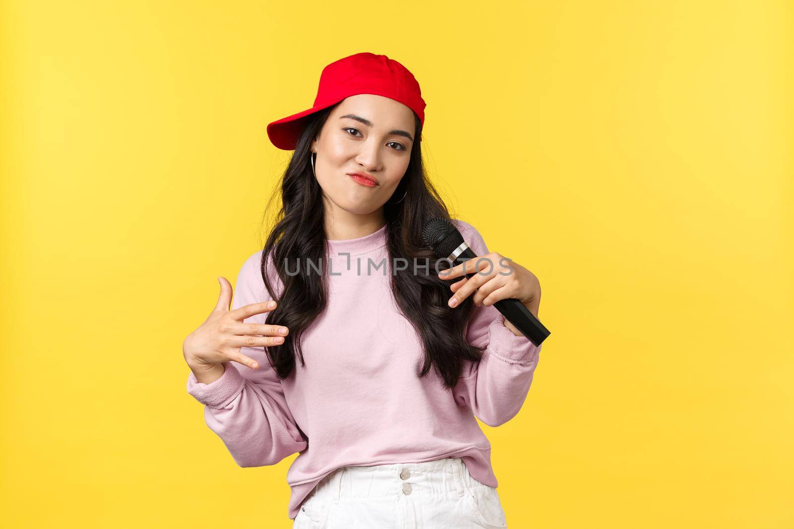 People emotions, lifestyle leisure and beauty concept. Stylish and cool young girl rapper in red cap, singing song and gesturing, performing with microphone, standing yellow background by Benzoix