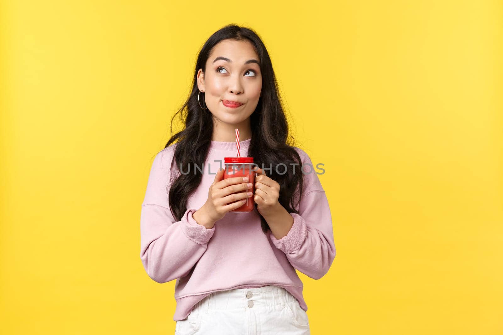People emotions, lifestyle leisure and beauty concept. Dreamy and thoughtful, cute asian girl searching inspiration, looking up and thinking while drinking smoothie over yellow background.