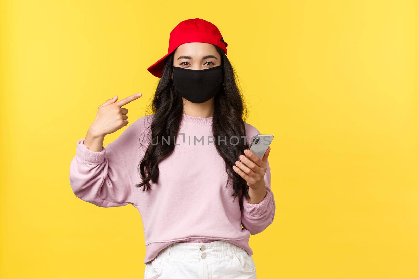 Covid-19, social-distancing lifestyle, prevent virus spread concept. Smiling cute asian girl pointing at face mask, asking protect health during coronavirus, holding mobile phone, yellow background.
