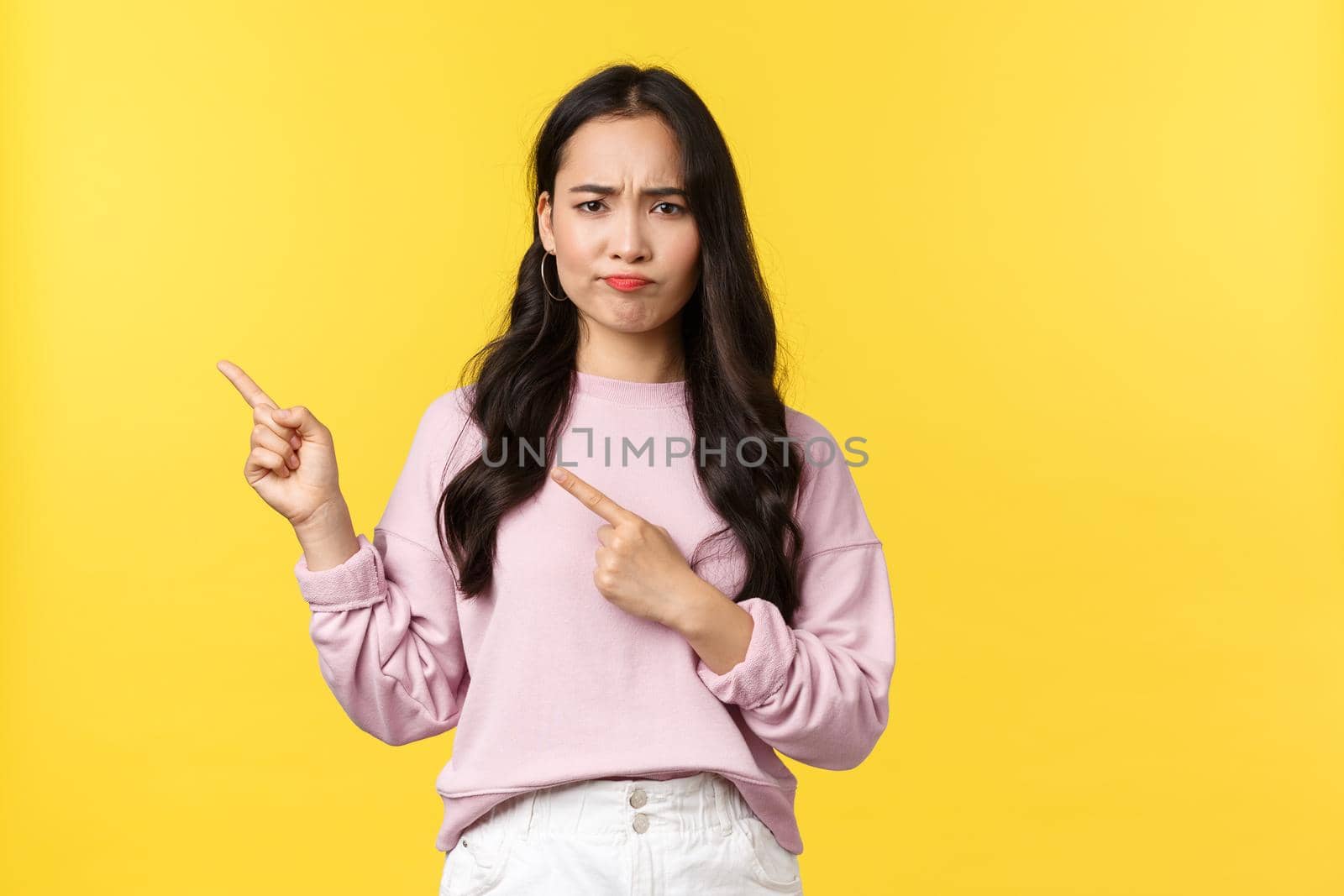 People emotions, lifestyle concept. Doubtful and indecisive cute asian girl looking puzzled, pouting and frowning as pointing upper left corner, asking question about banner, yellow background.