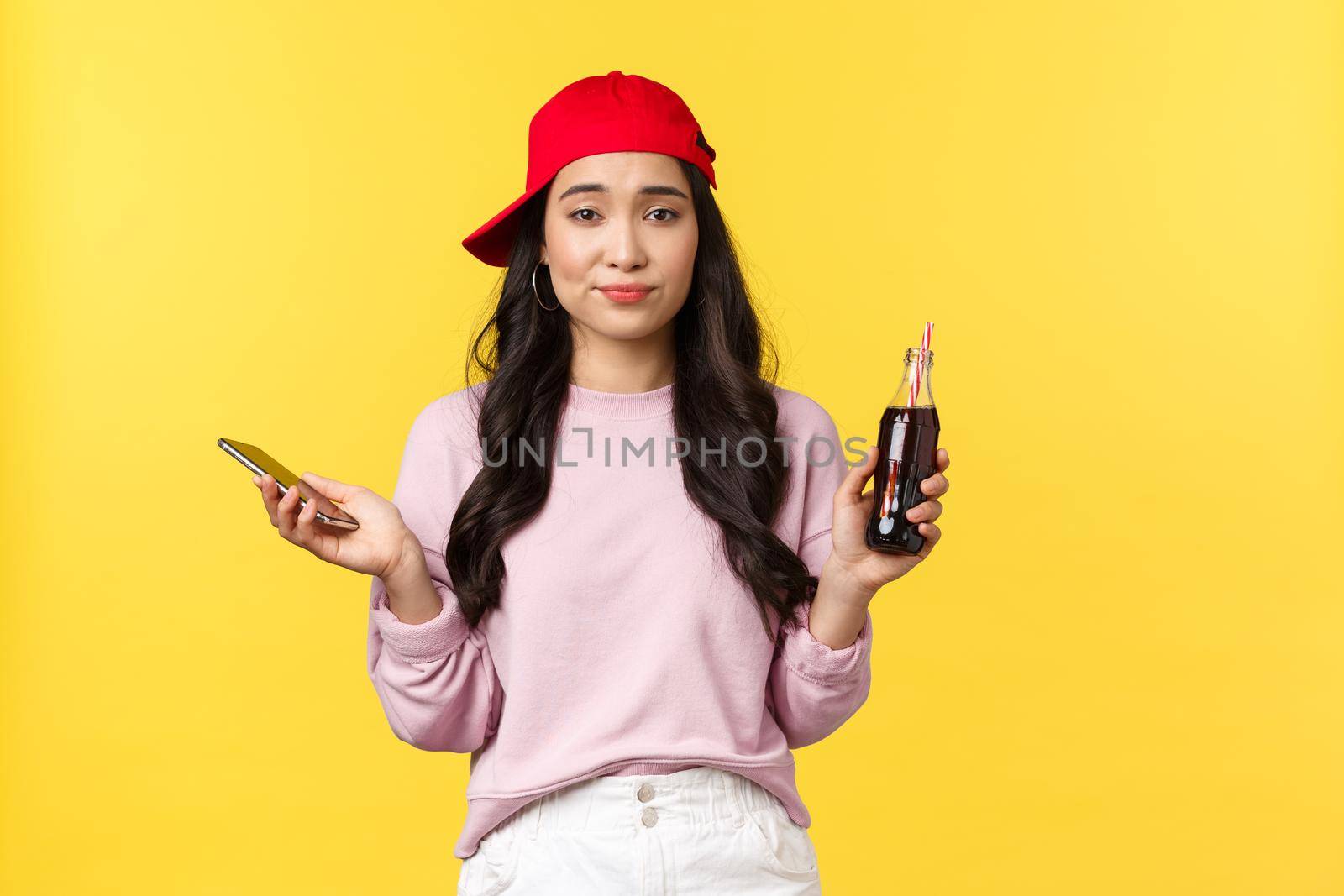 People emotions, drinks and summer leisure concept. Indecisive clueless cute asian girl in red cap, holding bottle with soda and mobile phone, shrugging unsure, yellow background.