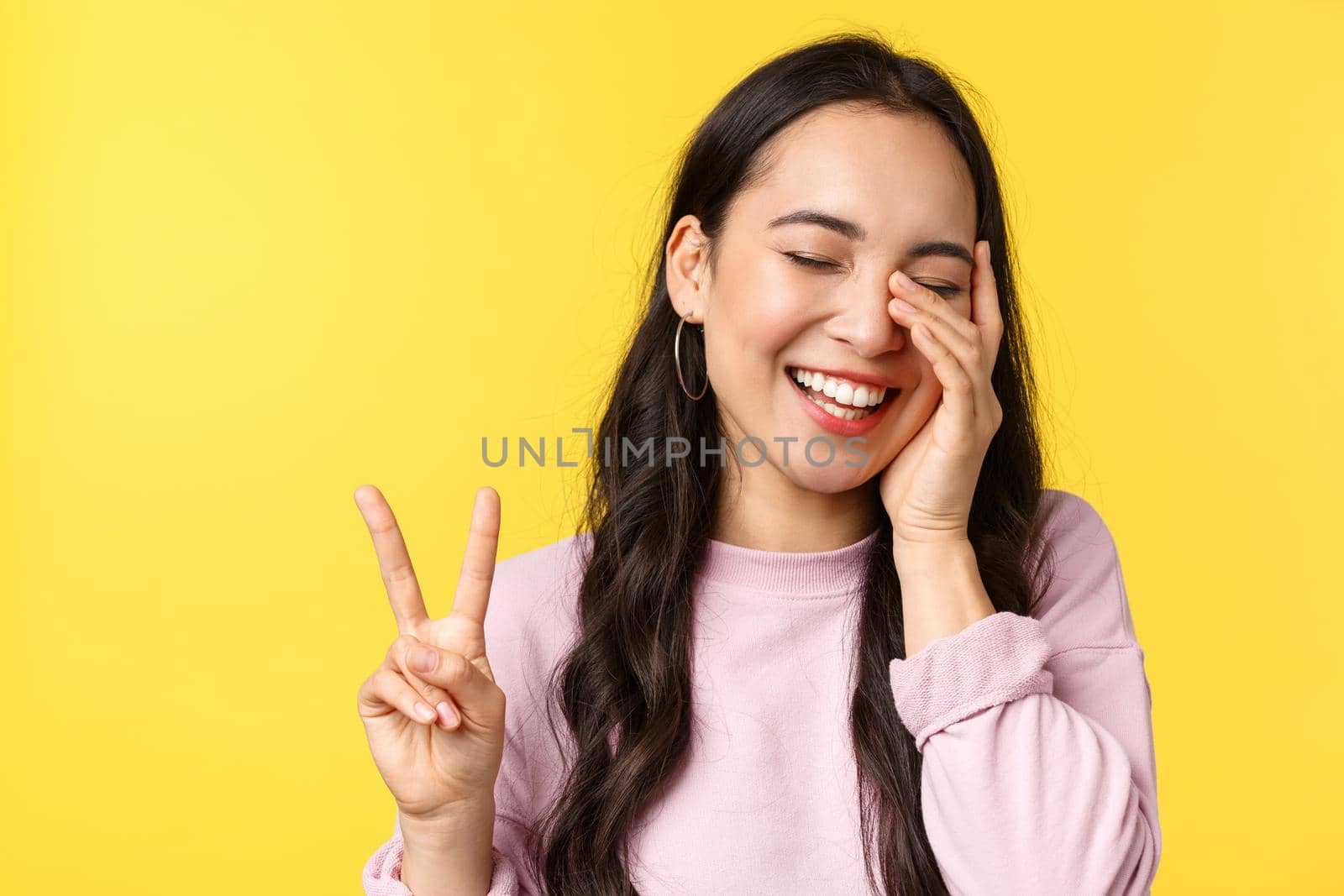 People emotions, lifestyle leisure and beauty concept. Close-up of carefree amused cute asian girl, showing peace sign touch face and laughing happy, standing yellow background by Benzoix