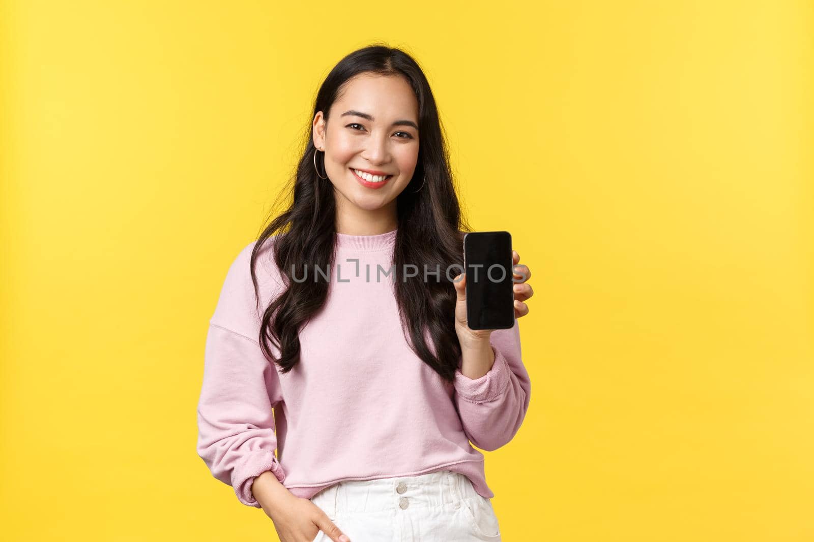 People emotions, lifestyle leisure and beauty concept. Smiling asian woman 20s, showing smartphone display, recommend application or mobile game, advertisement on screen, yellow background by Benzoix
