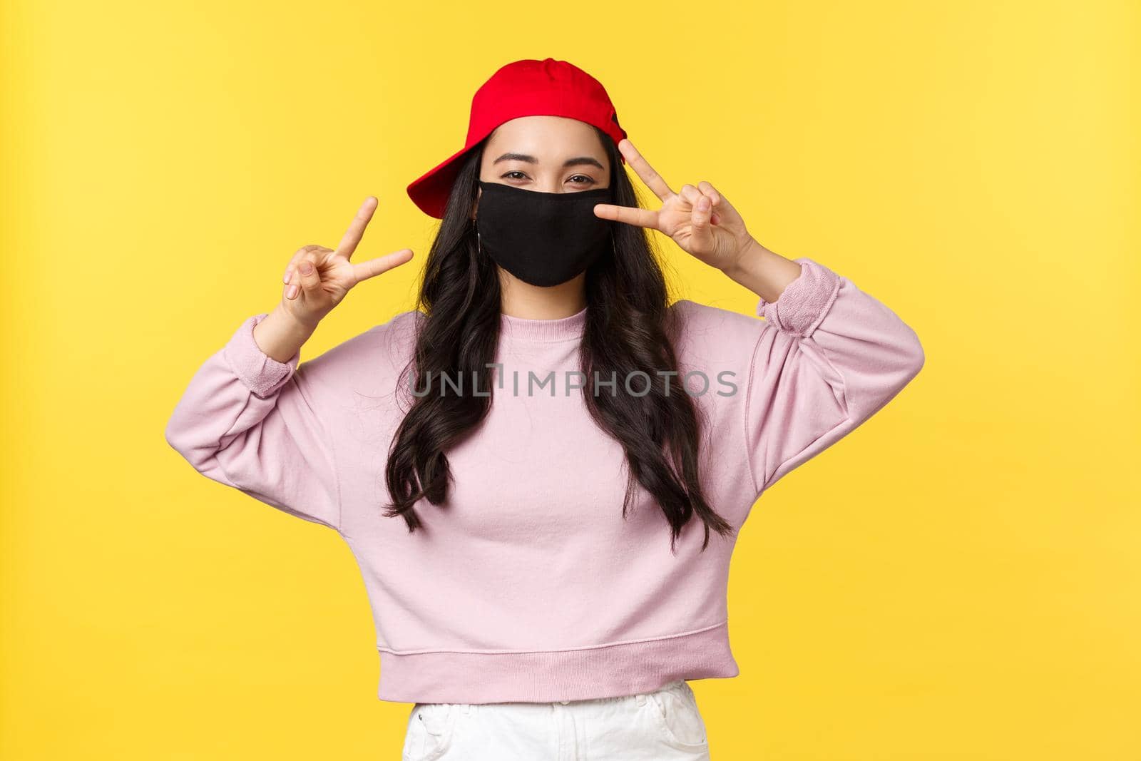 Covid-19, social-distancing lifestyle, prevent virus spread concept. Friendly cheerful asian girl in red cap and face mask, showing peace signs and smiling, standing yellow background by Benzoix