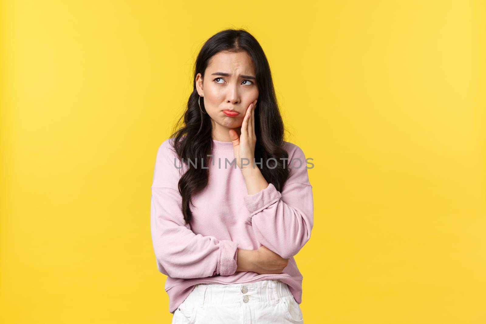 People emotions, lifestyle and fashion concept. Sad gloomy korean girl having toothache, looking upper left corner with upset grimace, touching cheek, standing yellow background.
