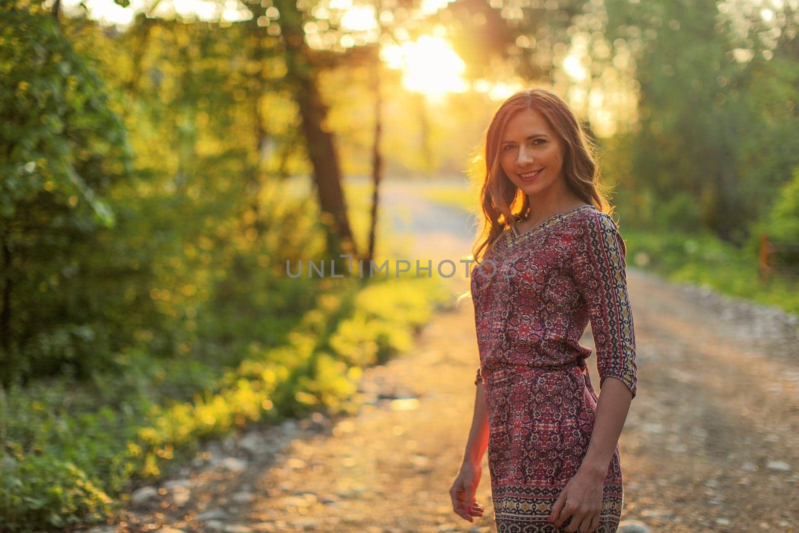 Young woman wearing dress, walking on forest path with golden sunset light in background.